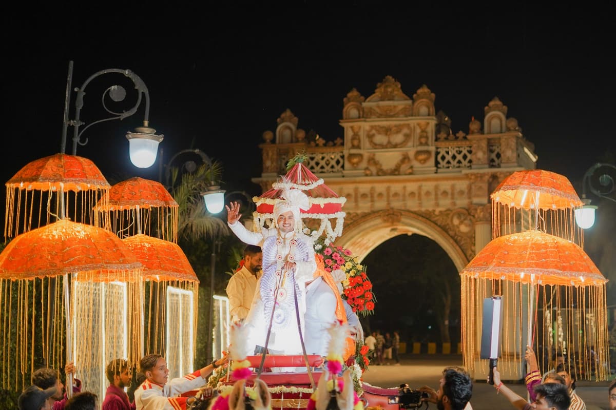 A night wedding procession: the groom stands on a decorated carriage under glowing orange fringed parasols, in front of a lit stone archway.