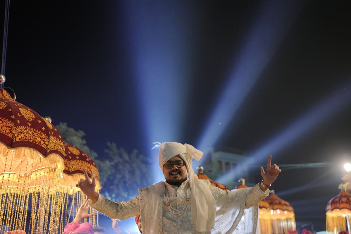A man in a white sherwani and turban dances with arms raised beside fringed gold parasols, with searchlight beams sweeping the night sky.