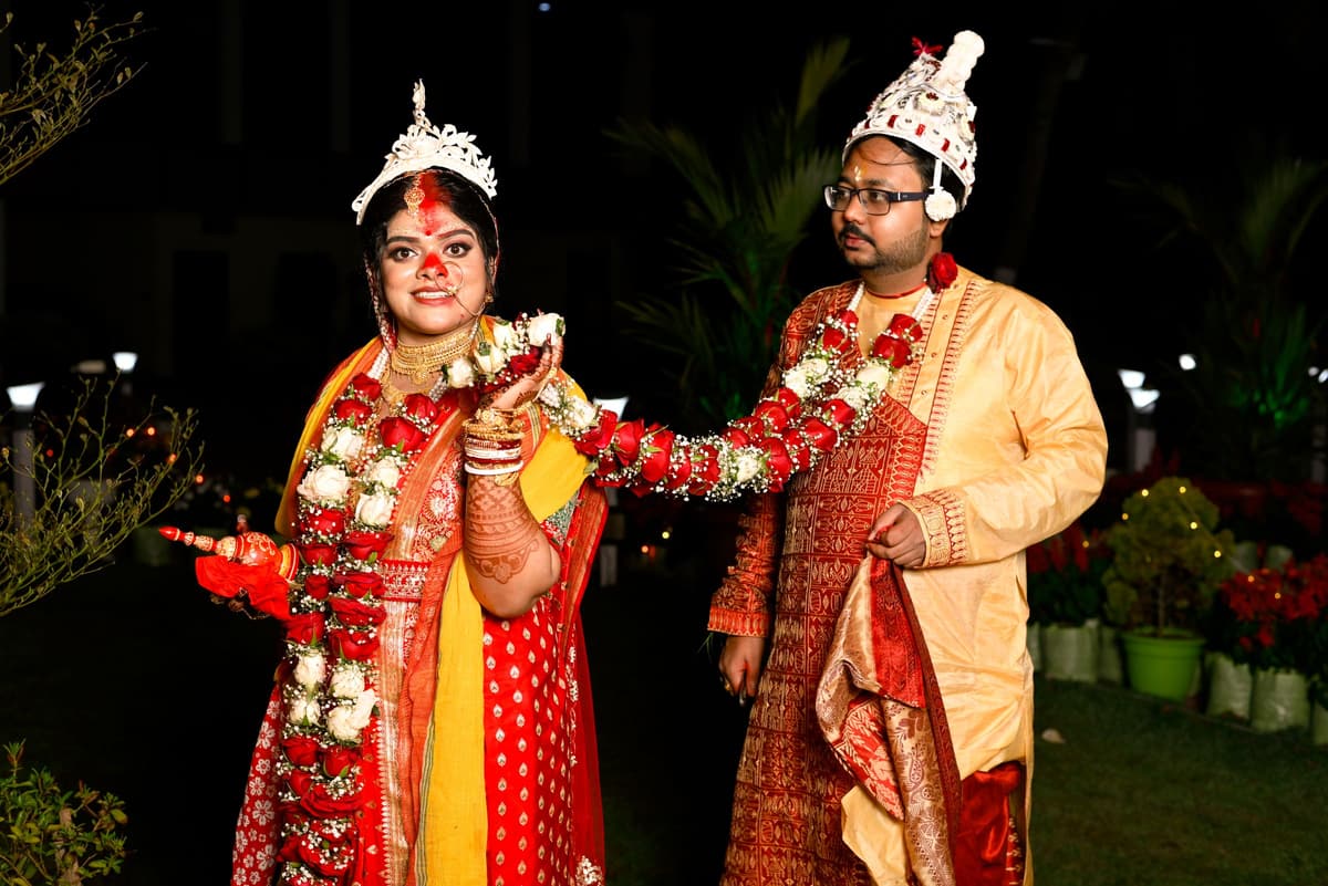 A Bengali couple at night in white shola topor and mukut, red-and-white saree, heavy flower garlands; one holds a lit diya.