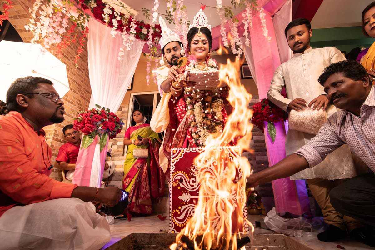 At a Bengali wedding a tall fire blazes in the foreground; the couple in topor and mukut stand behind it while relatives make offerings.