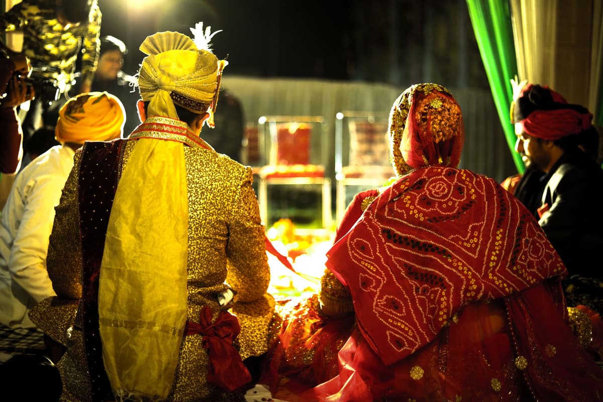 Seen from behind at night: the couple sit at the fire, one in a gold sherwani and yellow safa, the other in a red-and-white bandhani odhni, with relatives in turbans nearby.