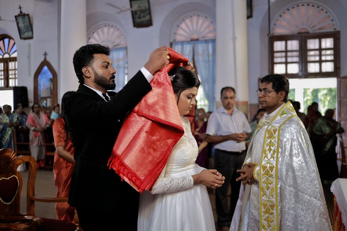 Inside a Kerala church, a man in a dark suit drapes a red manthrakodi saree over the head of the woman beside him while a priest in gold vestments looks on.