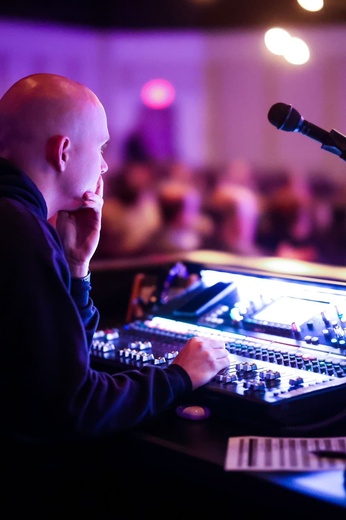 An audio technician at an illuminated mixing console during a ballroom event, face lit by the desk, the crowd blurred behind in purple uplight.