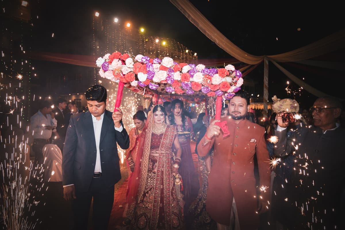 At night, relatives carry a canopy of roses over a woman in a red lehenga as she walks in, cold sparks fountaining on both sides.