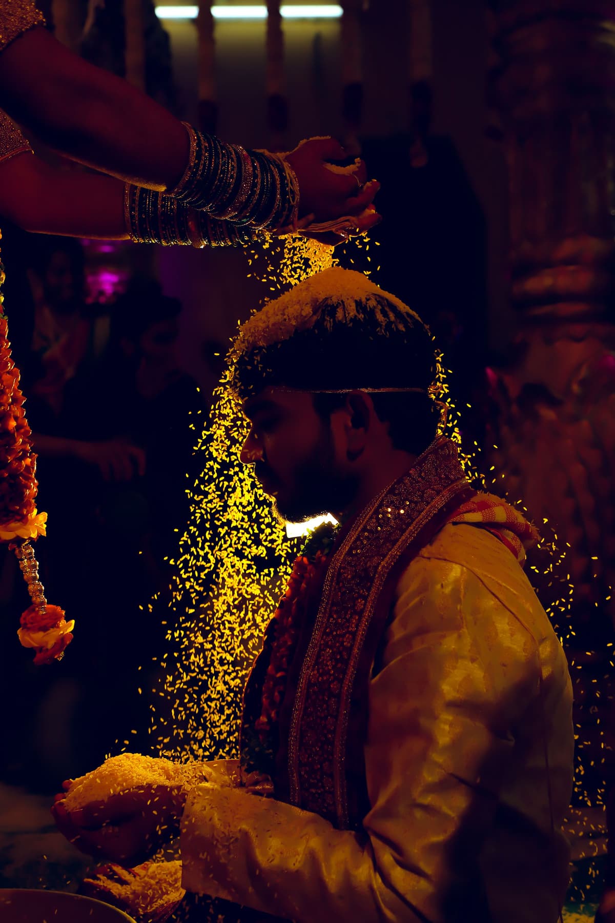 In warm darkness, golden rice grains fall in a glittering stream over a man in a silk kurta with a garland.