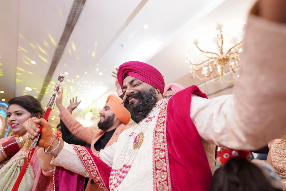 A laughing man in a pink turban and embroidered shawl dances with relatives under chandeliers as sparkles fall.