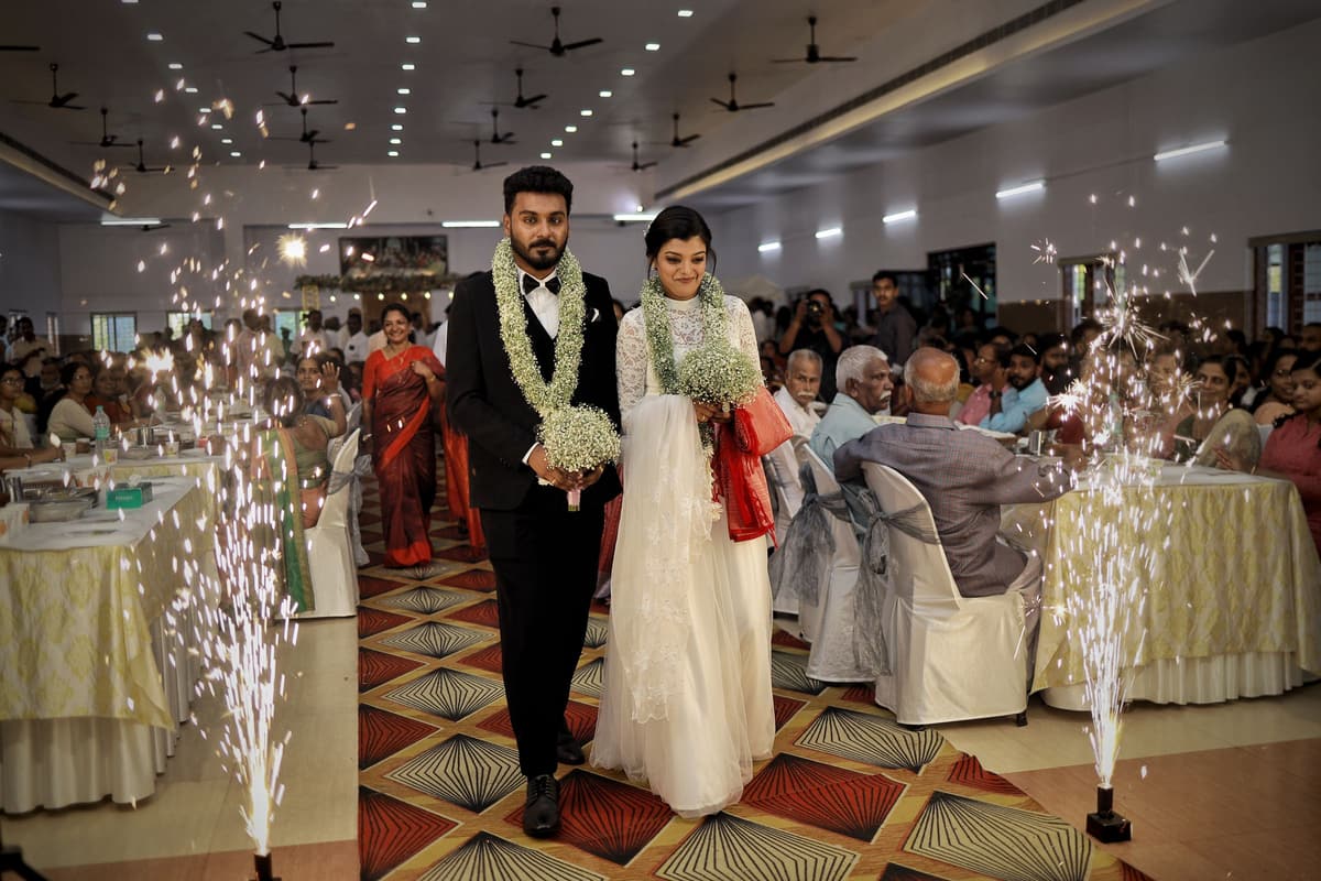 A garlanded couple walk into a reception hall between cold-spark fountains, one carrying a red manthrakodi saree over the arm, guests in sarees at tables.