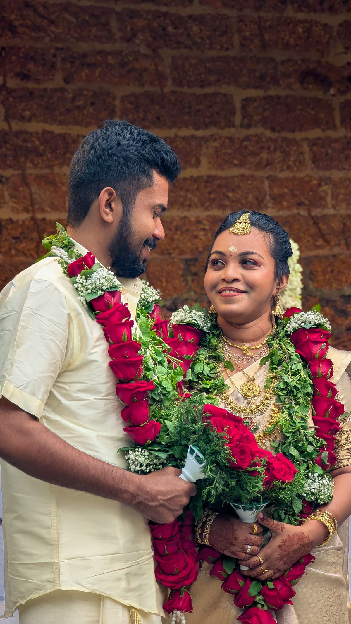 A Malayali couple in white-and-gold kasavu, heavily garlanded in red and green, smile at each other against a laterite stone wall.