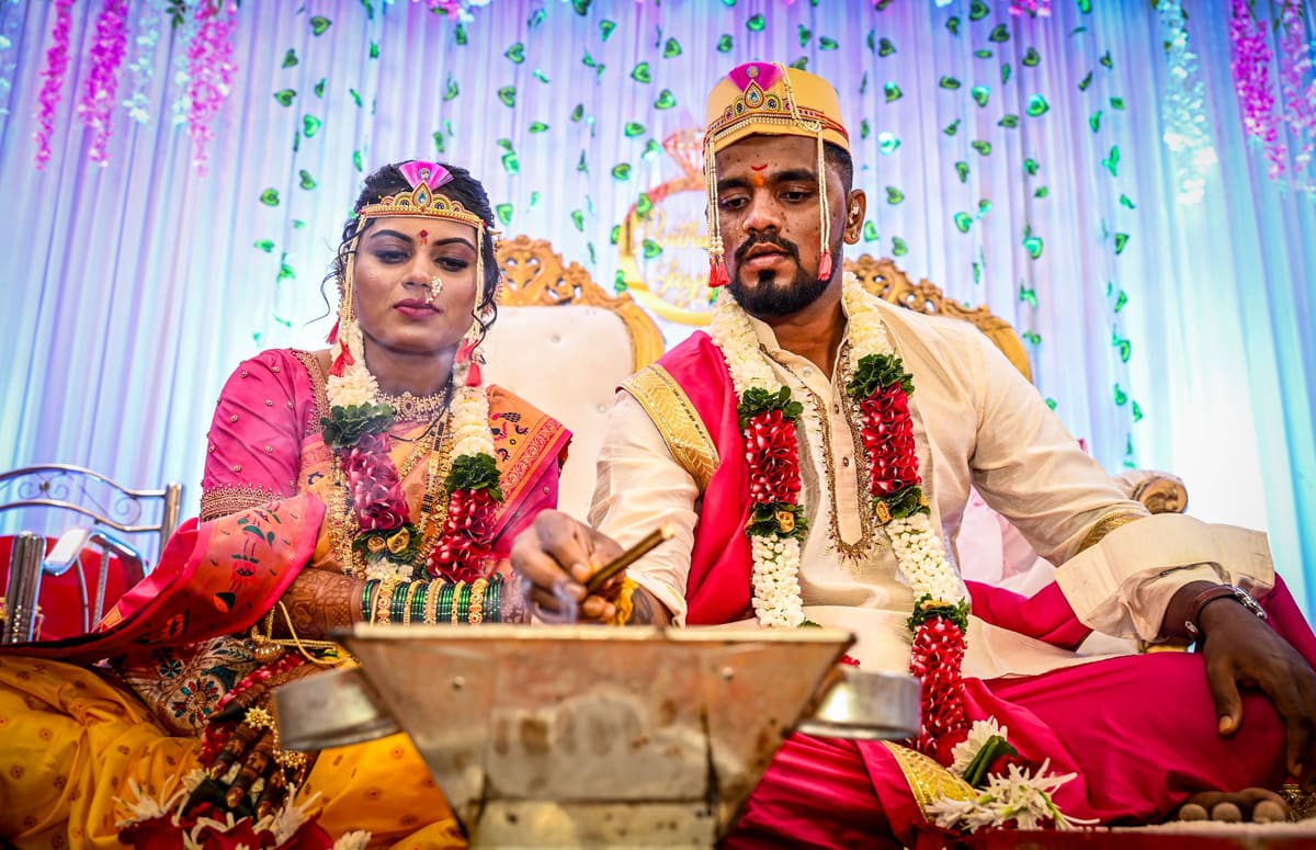 A Maharashtrian couple seated at the havan, both wearing pearl mundavalya across their foreheads, garlanded, with pastel drapes behind.