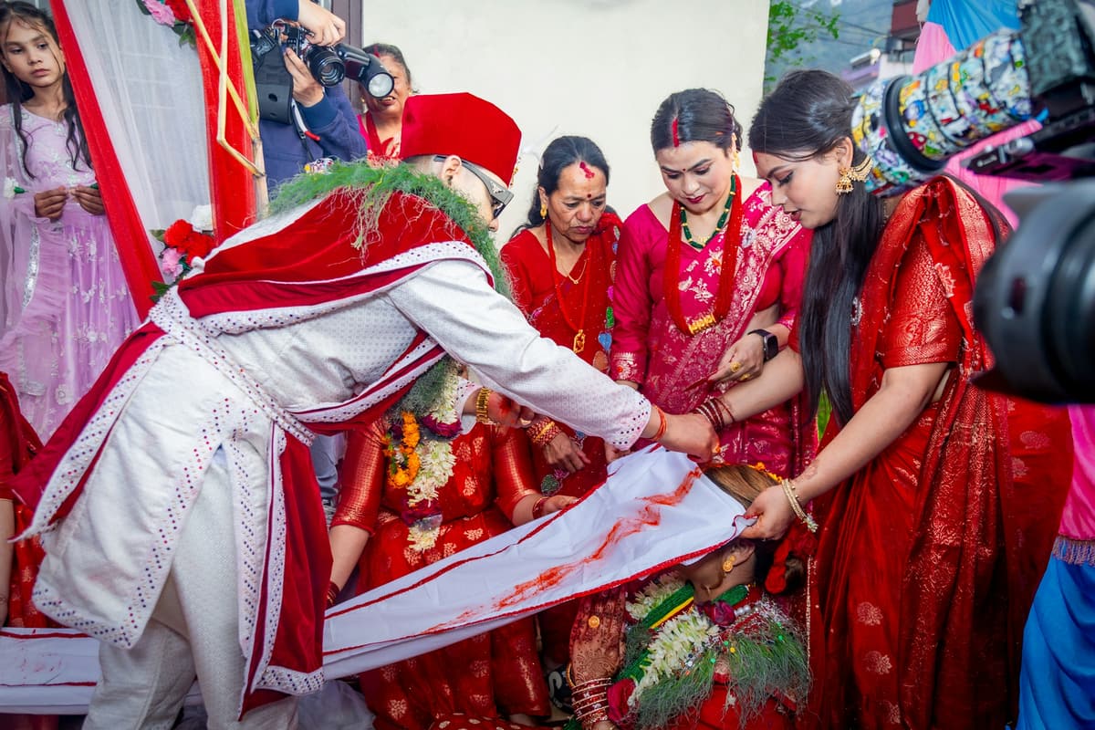 At a Nepali wedding a man in a red-and-white daura suruwal draws sindur along a white cloth over the bowed head of the woman in red, relatives crowding close.