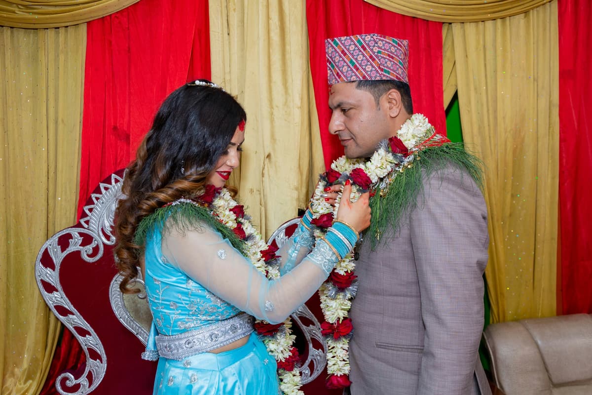 A couple exchange garlands at a Nepali engagement, the man in a dhaka topi, in front of red and gold drapes.