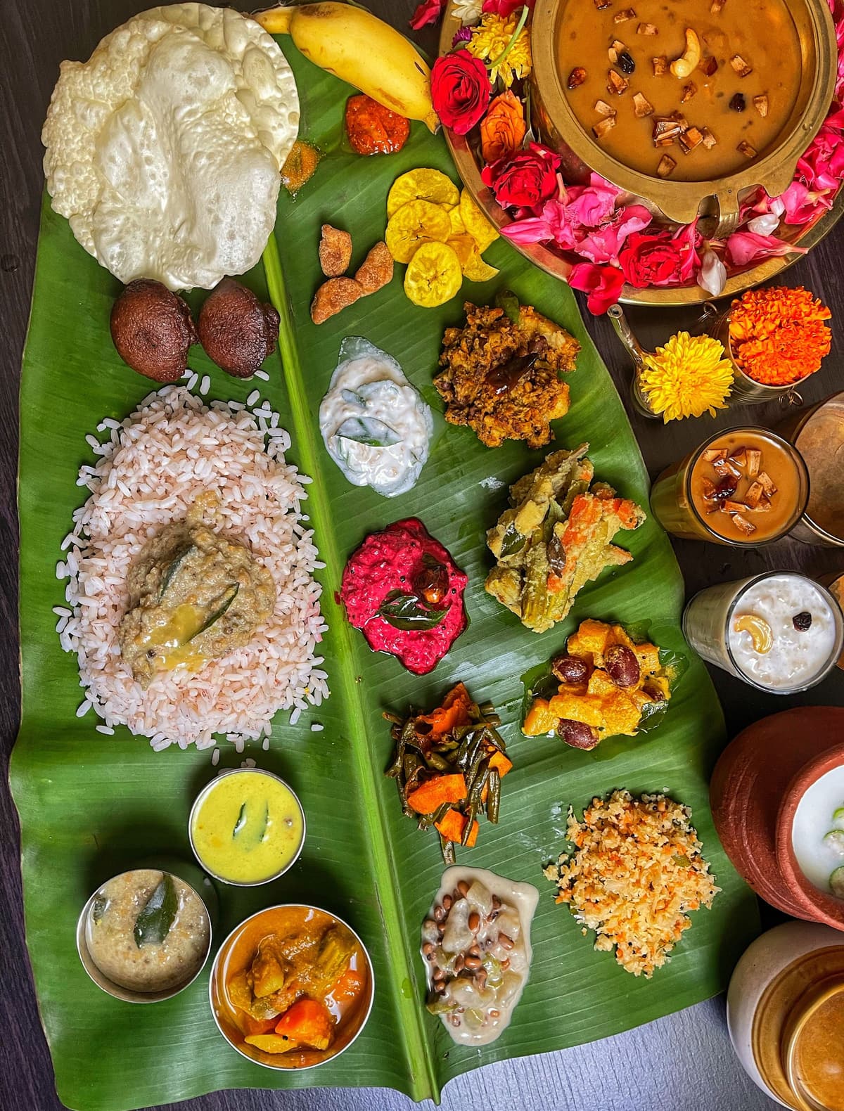 Top-down view of a full banana-leaf meal: rice, a dozen curries and sides, papadum, banana, payasam and marigold petals.
