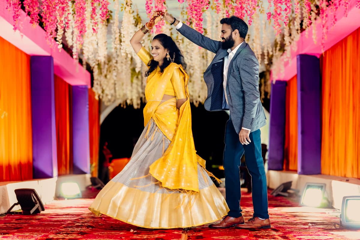 A couple dances on a stage dressed in orange and purple drapes under hanging flowers, with LED uplights on the stage floor.