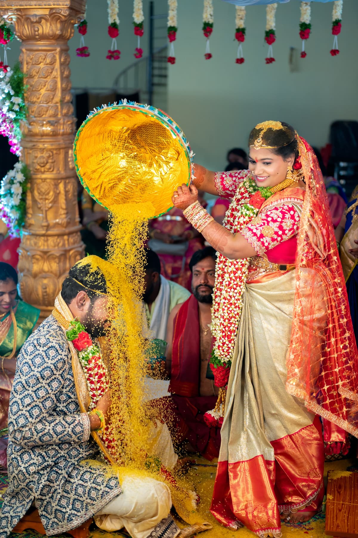 On a pillared mandapam, a woman in a red-and-gold pattu saree with jasmine pours a stream of turmeric rice from a gold bowl over the seated man.