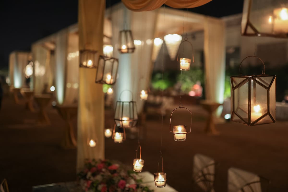 An outdoor evening venue hung with brass candle lanterns, warm glow on draped pillars and lounge seating.