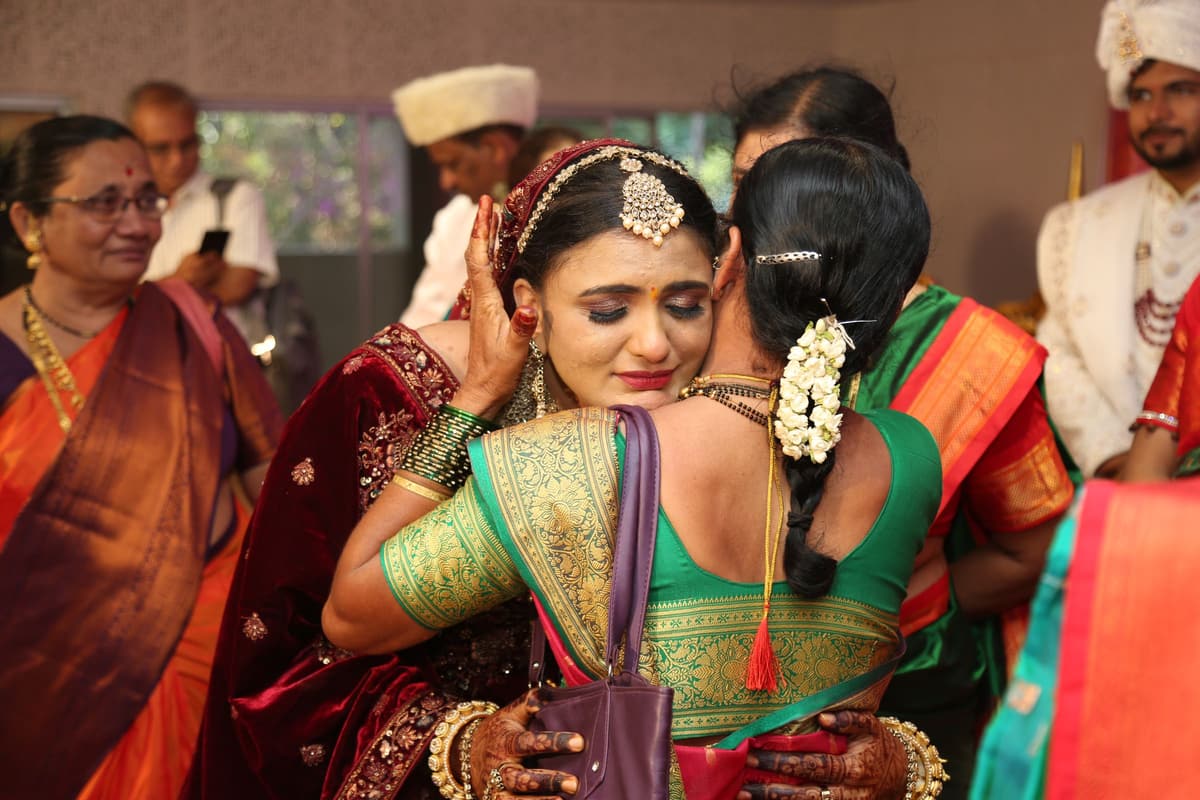 A tearful woman in bridal jewellery embraces a relative in a green silk saree while older relatives, one in a white cap, look on.